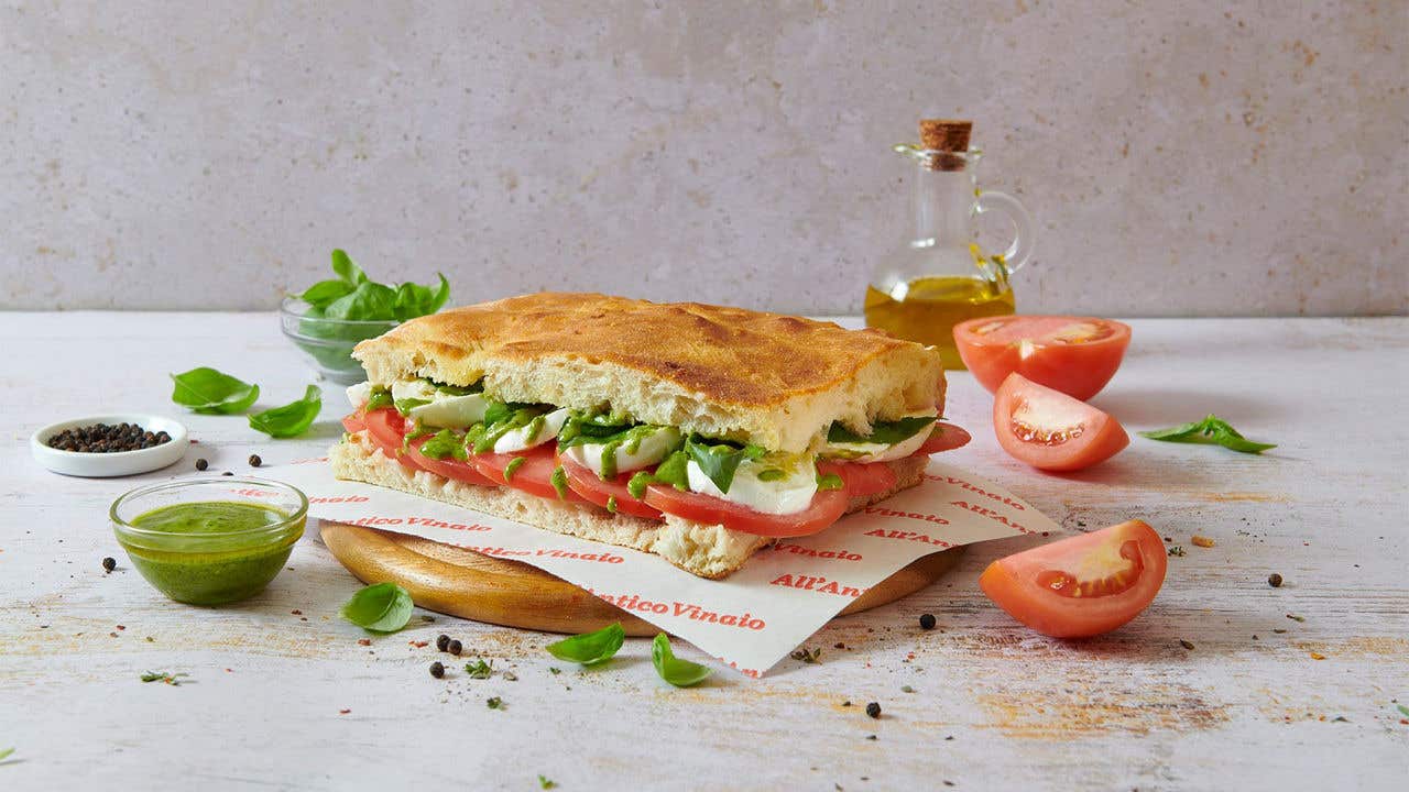 Panini sandwich with tomato, lettuce and mozzarella cheese on a round wood plate surrounded by slices of tomato, seasoning and a bottle of olive oil. 