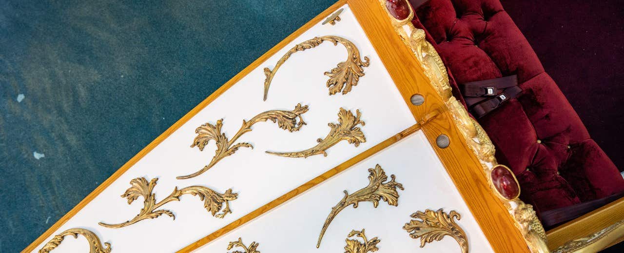 Close-up of an ornate table with golden floral designs and a red cushioned seat in the background.