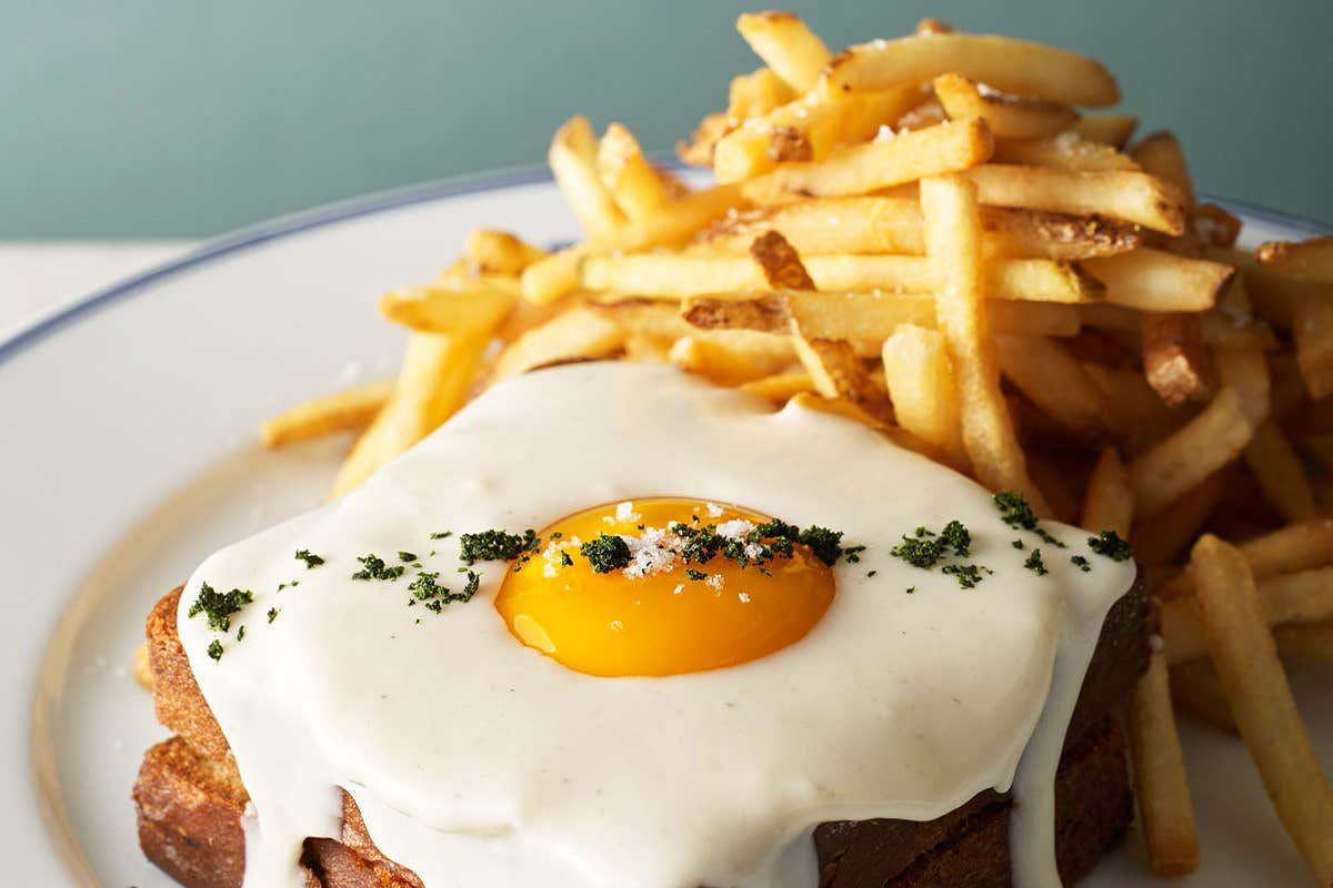 A dish with a sunny-side-up egg over toast, covered in white sauce, with a side of crispy French fries on a white plate.