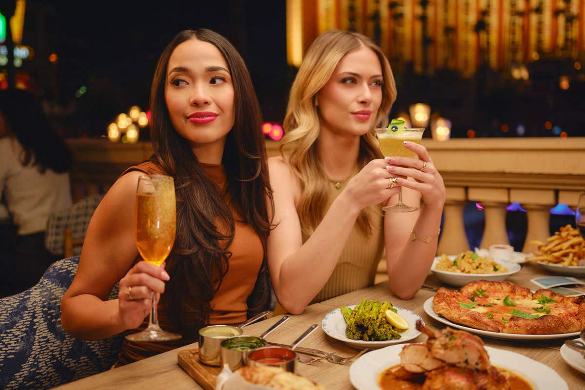Two girls sitting at a table with an assortment of food in front of them while holding their drinks. 