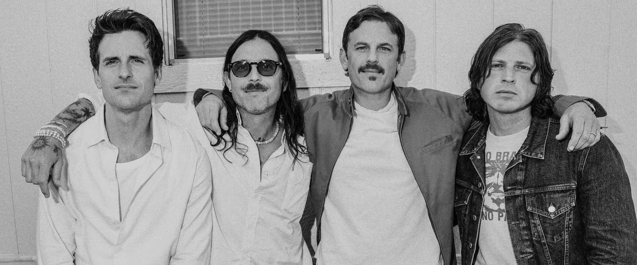 A grayscale photo of Kings of Leon band members standing together in front of a building looking directly at the camera. 