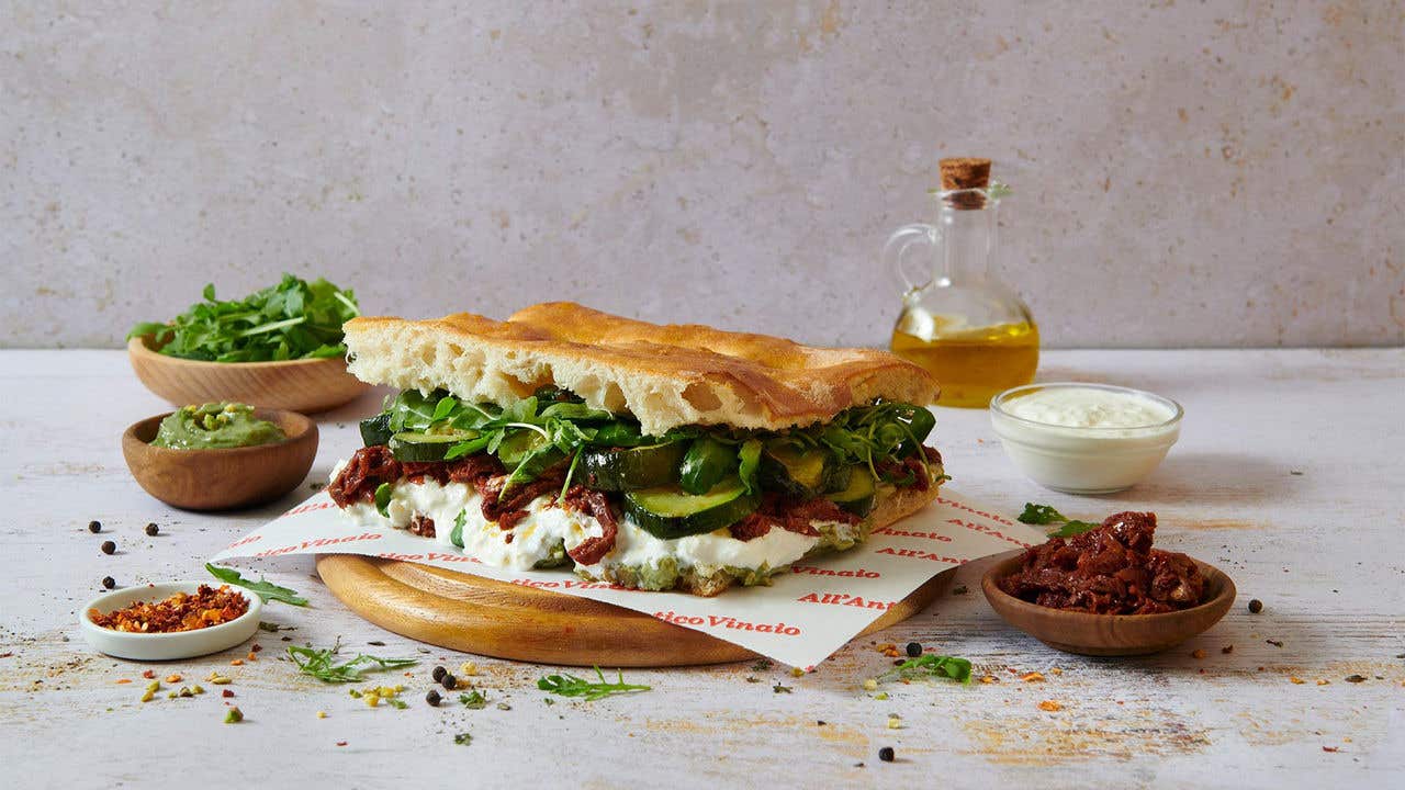 Panini sandwich with cream cheese, pesto, lettuce and cucumbers on a round wood plate surrounded by different seasonings. 