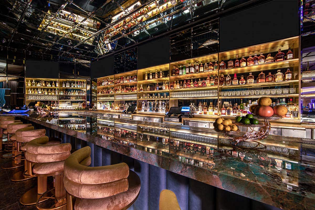 A reflective bartop and ceiling with a golden lit arrangement of bottles on the shelves in the background