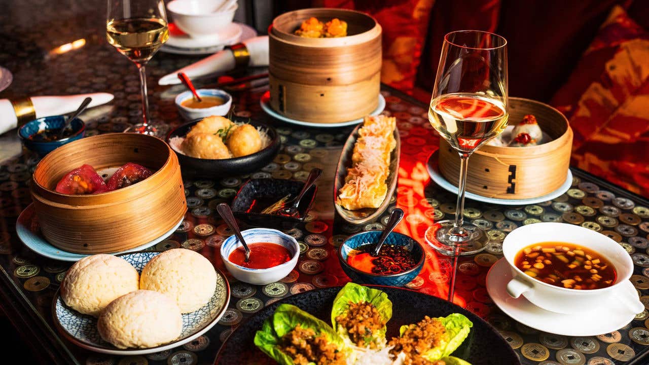 Table spread with dim sum, soups, buns, lettuce wraps, and glasses of wine.