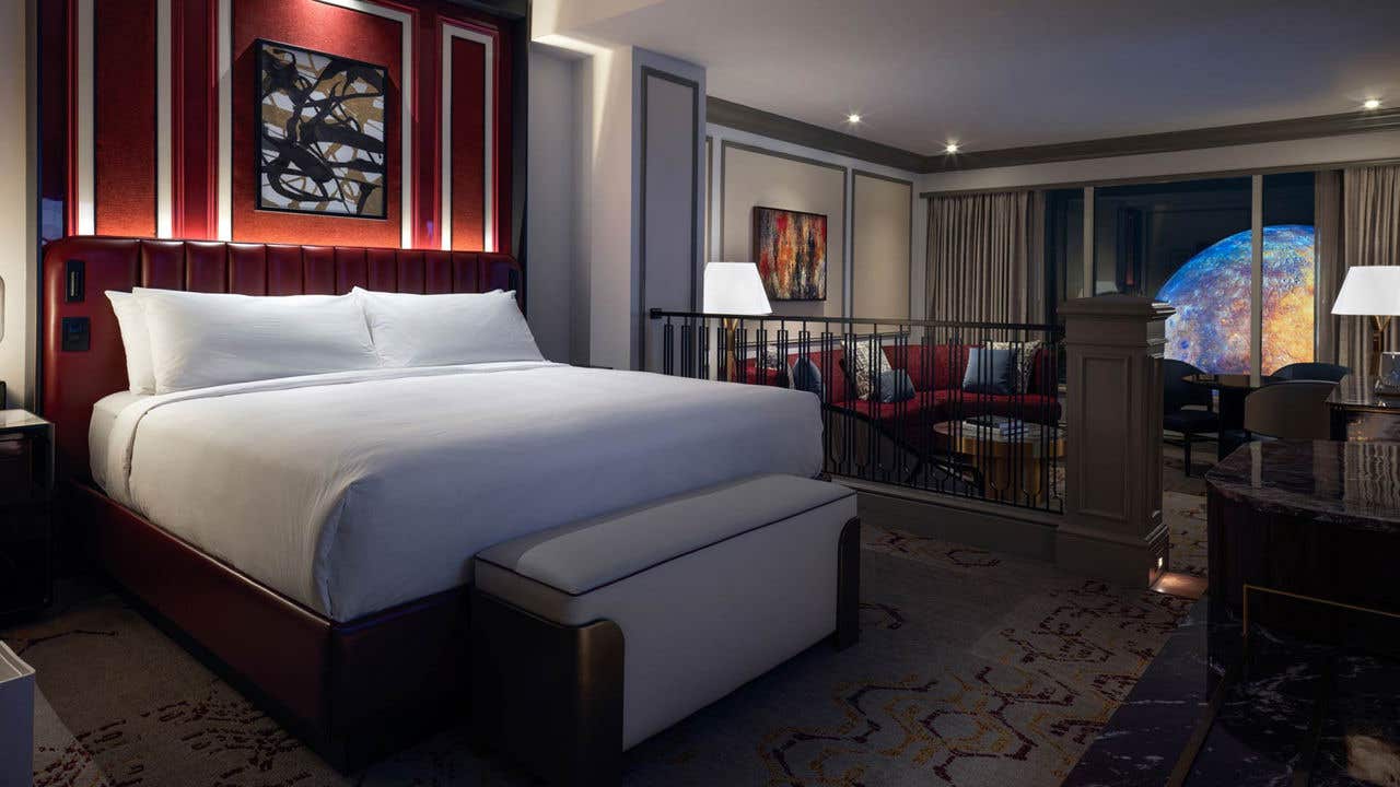 A hotel room at night with a king bed on the left, a living room area on the right, and a view of the Sphere through window on the right.