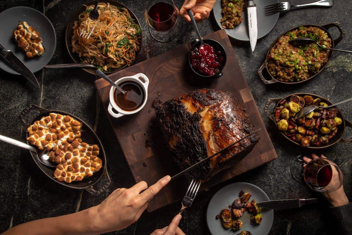 Piece of meat being carved surrounded by side dishes like burnt marshmallows, brussels sprouts and cranberries. 