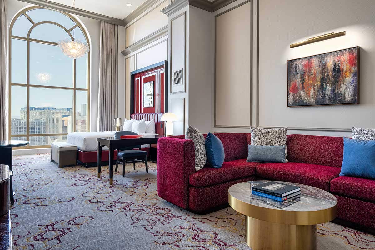 A hotel room with a king bed, desk with a chair, curved red couch, and a gold coffee table. 